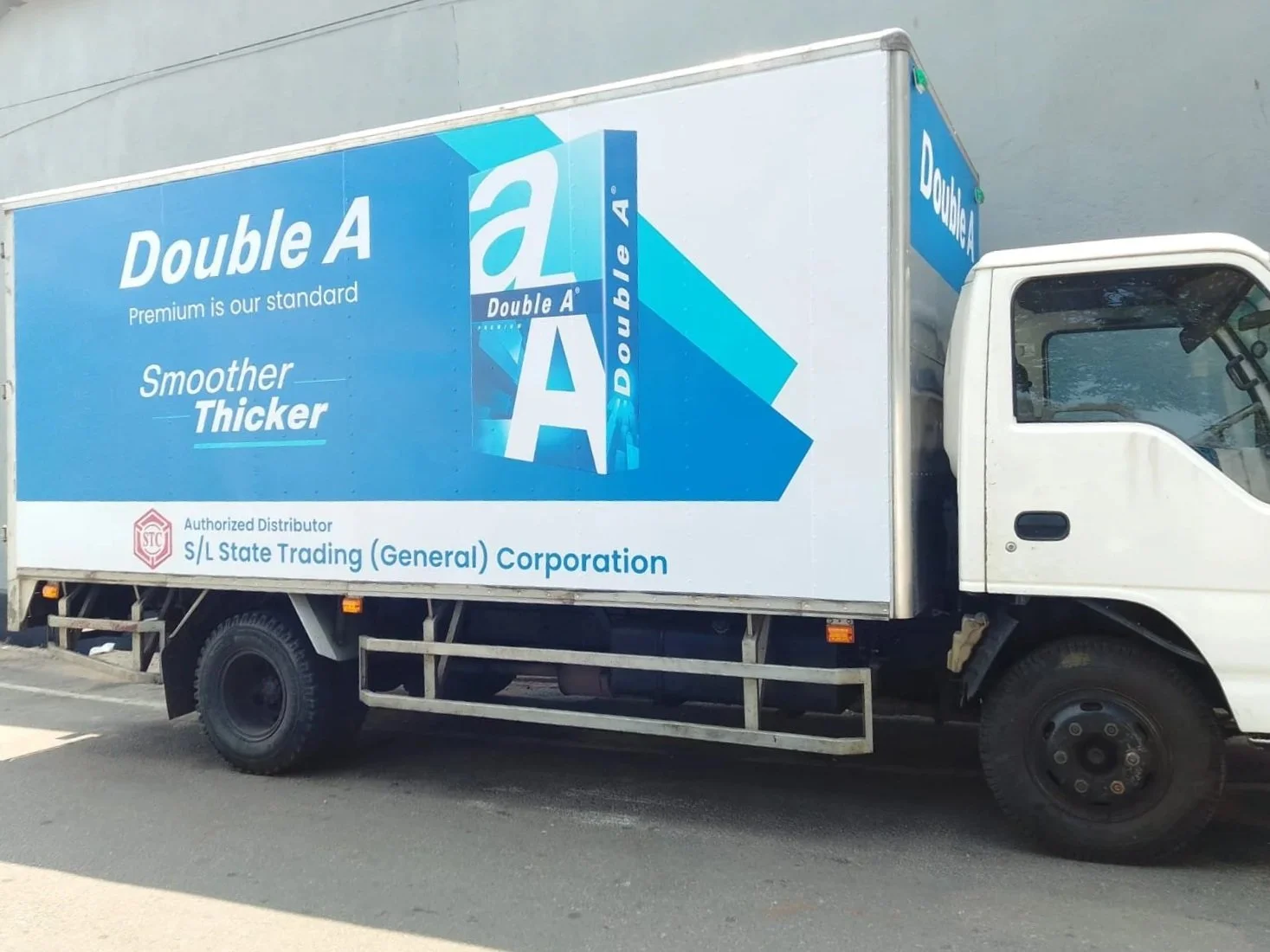 A delivery truck with an advertisement for Double A brand paper products on its side. Text includes "Double A," "Premium is our standard," "Smoother Thicker," and mentions an authorized distributor, S/L State Trading (General) Corporation.