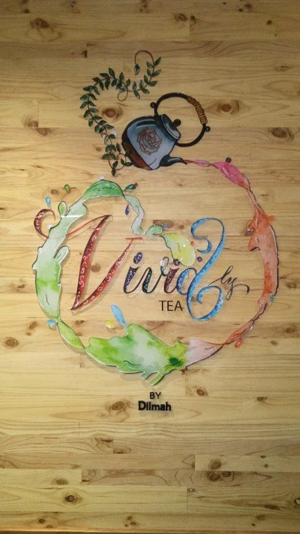 Colorful sign with the word "Vivid" and a teapot pouring tea, surrounded by a watercolor floral circle, on a wooden wall.
