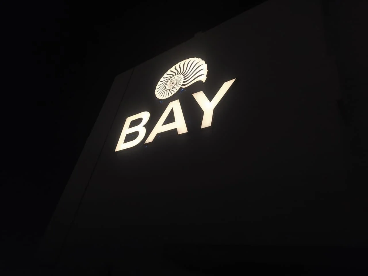 A illuminated sign with a stylized shell logo and the word "BAY" in large letters on a dark background.