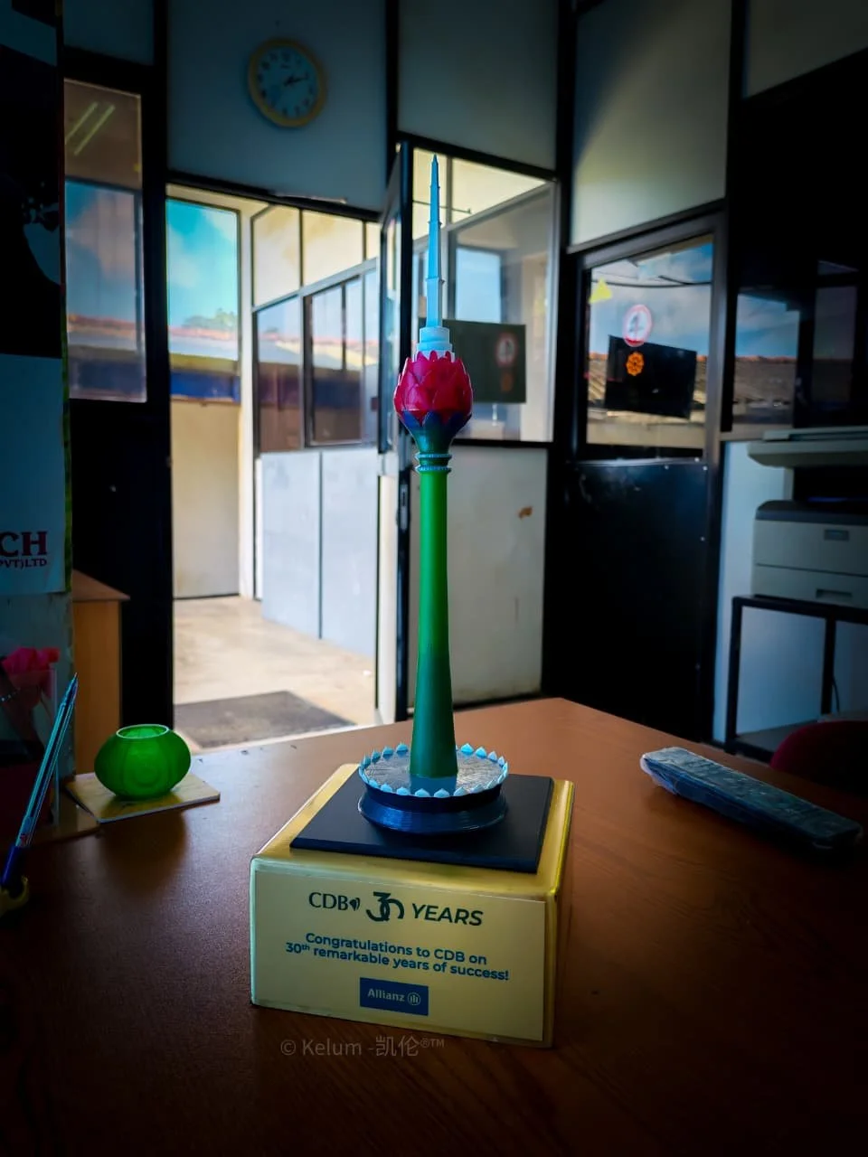 A model of the Thitron Tower placed on a yellow box inside an office, with an open door and windows in the background showing a blue sky and a clock on the wall.