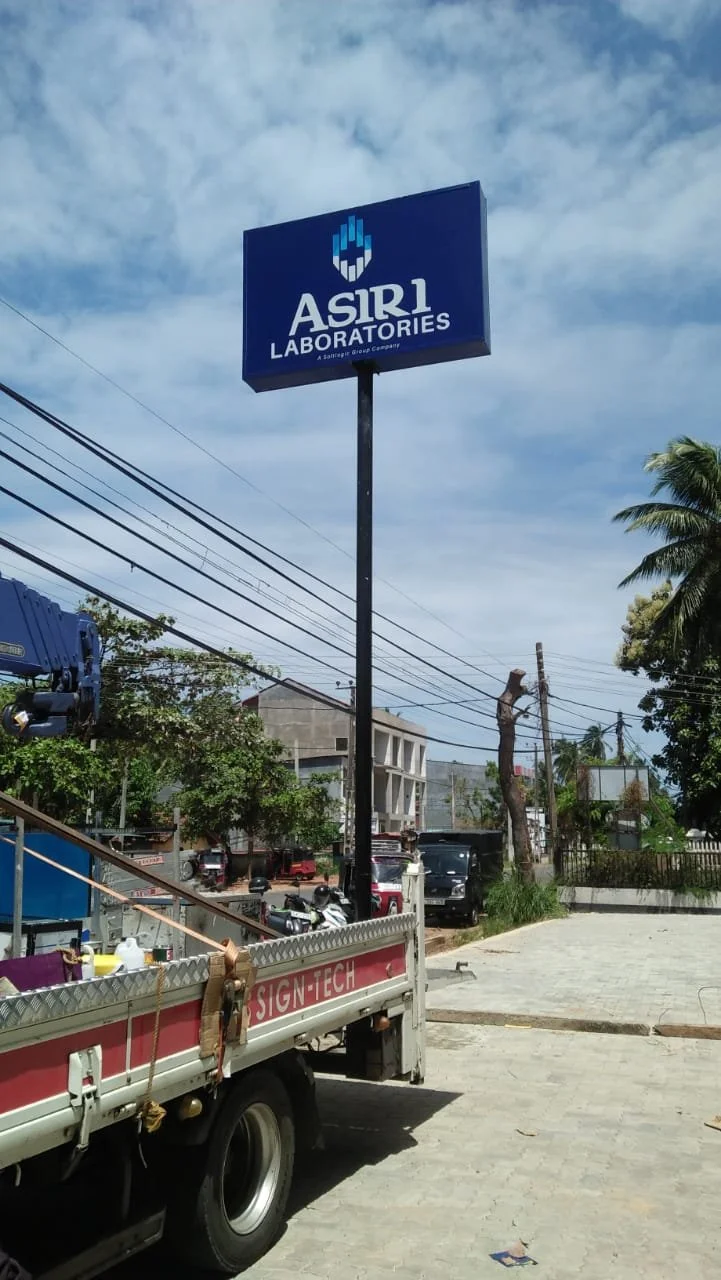 A tall sign for ASIR1 Laboratories with a blue background and white text, situated on a black pole outdoors on a partly cloudy day. There are trees, power lines, a warehouse-type building, parked cars, and a truck nearby.