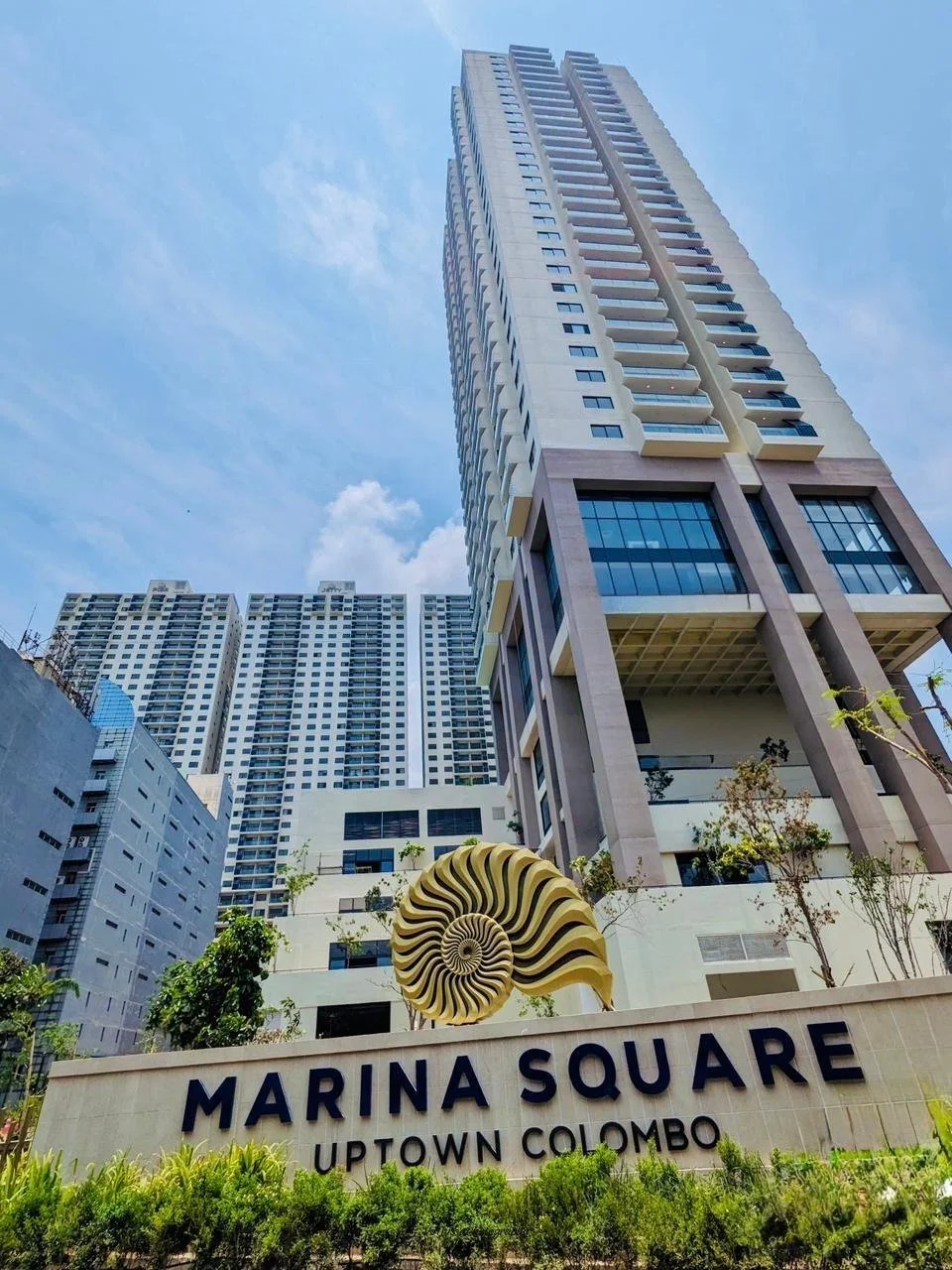 A modern high-rise building with a large sign reading 'Marina Square Uptown Colombo' at its base, surrounded by trees and adjacent buildings, under a clear blue sky.