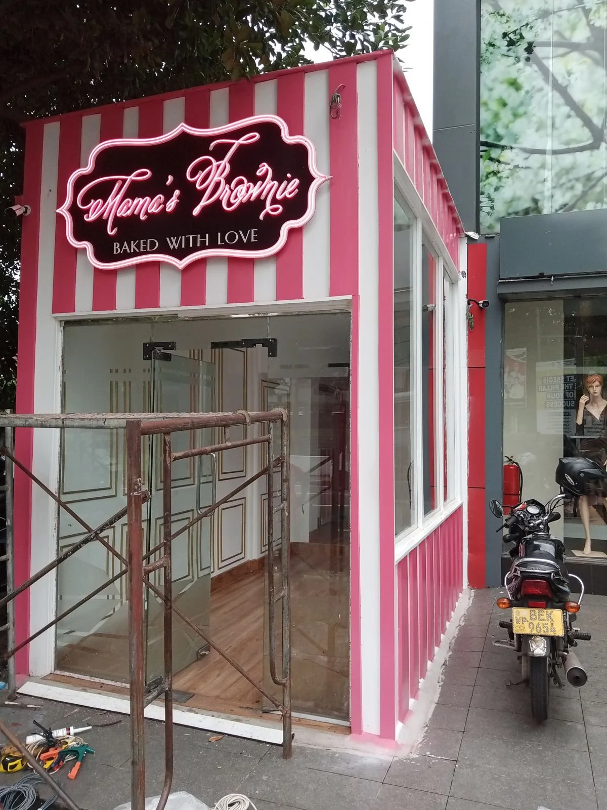 A small storefront with pink and white vertical stripes, a neon sign that reads "Mama's Promie" with a slogan "Baked with Love" beneath, and large glass windows. There are construction tools, scaffolding, and motorcycles outside.