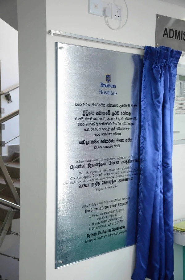 A plaque or signboard at Browns Hospital, with some text in Sinhalese and English, partially obscured by a blue curtain, mounted on a wall near a staircase and an electrical outlet.