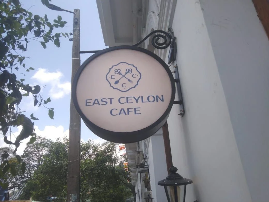 A round sign for East Ceylon Cafe hanging outside on a black metal bracket with decorative scrollwork, against a partly cloudy sky and some trees.