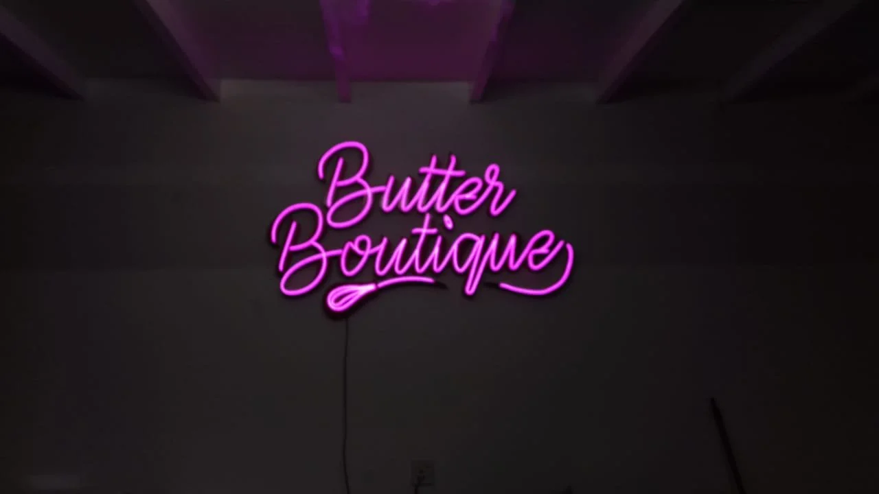 Pink neon sign reading 'Butter Boutique' on a gray wall.