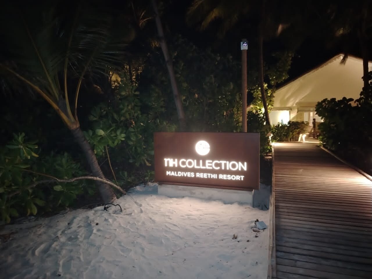 Illuminated sign at Maldives Reethi Resort, indicating 'The Collection' on a sandy walkway at night surrounded by foliage.