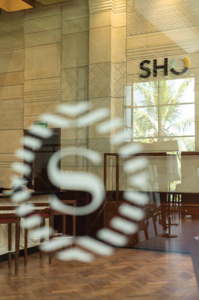 Interior view of a store called SHO with a large window showing palm trees outside, and a blurred logo or sign in the foreground.