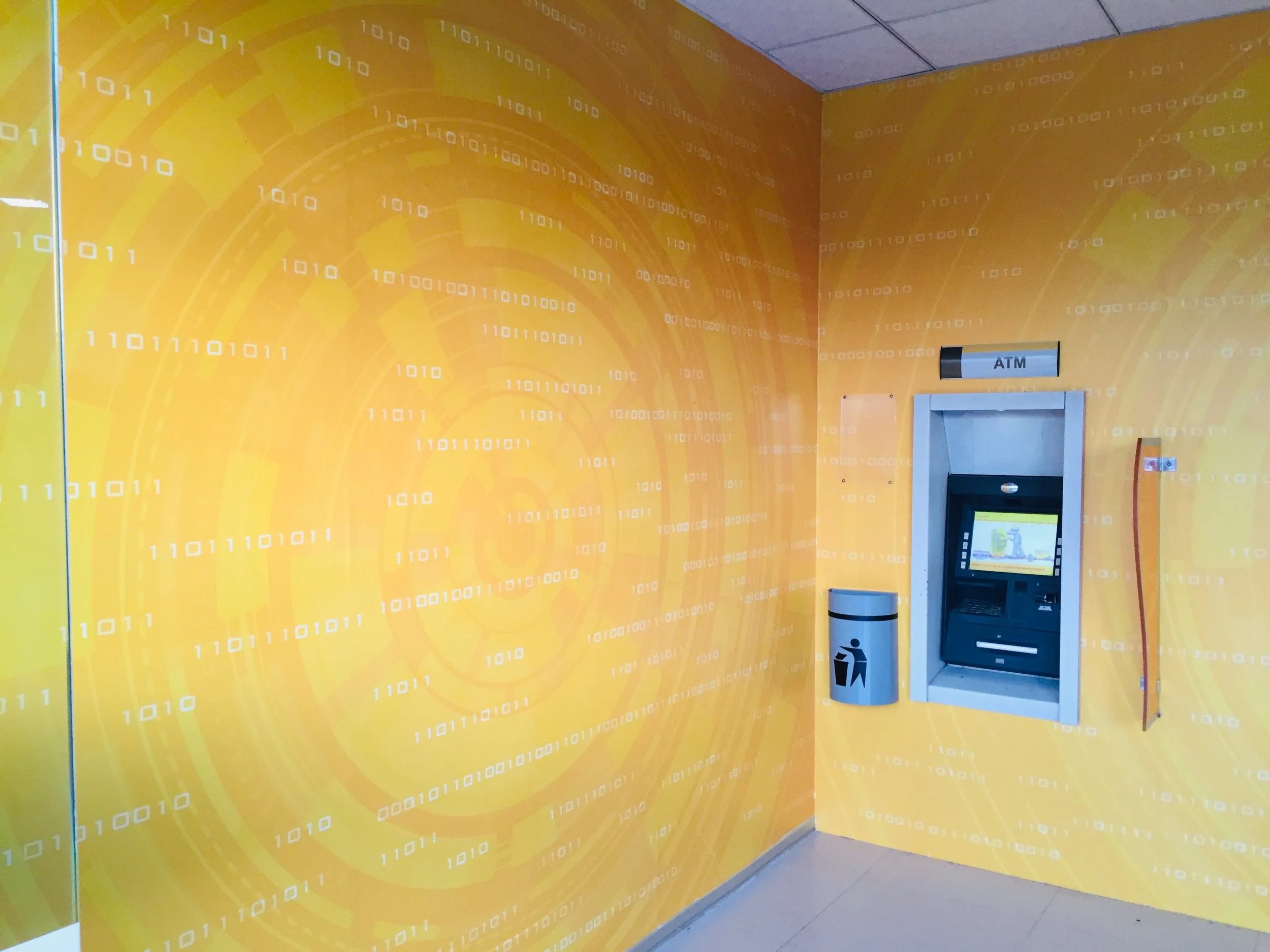 The image shows a brightly colored yellow and orange wall with binary code patterns, featuring a digital ATM machine in the corner with a screen and a card slot, and a trash bin nearby.