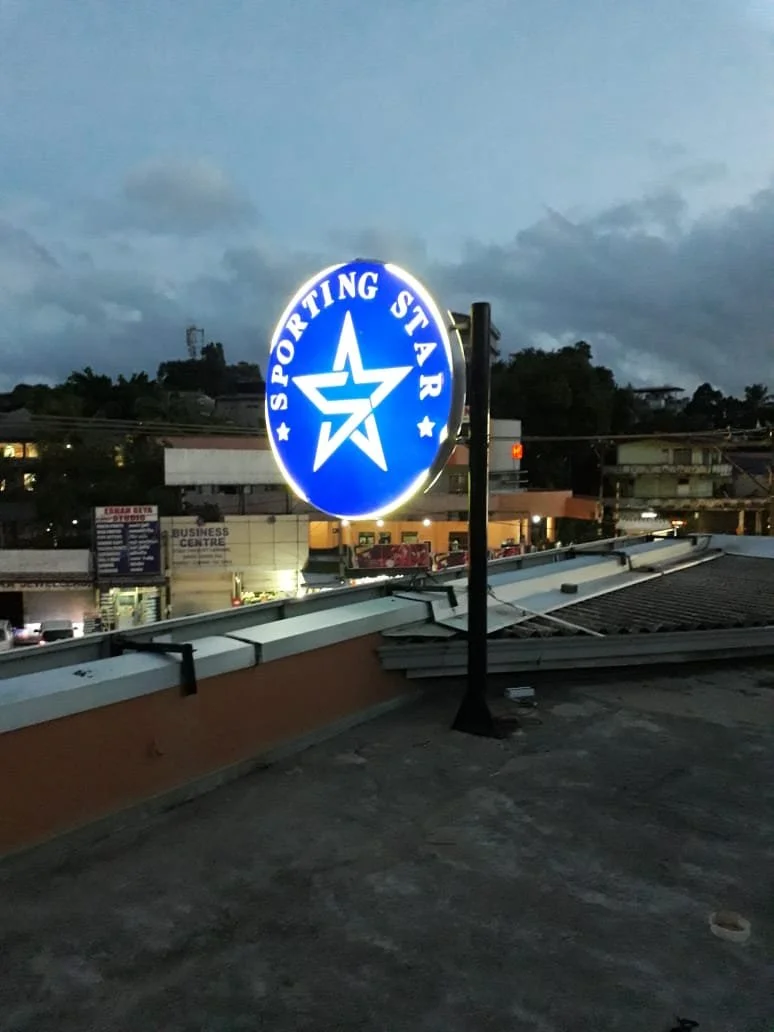 Illuminated blue and white round sign with the text 'SPORTING STAR' and a star logo, mounted on a pole on a rooftop against a cloudy evening sky.