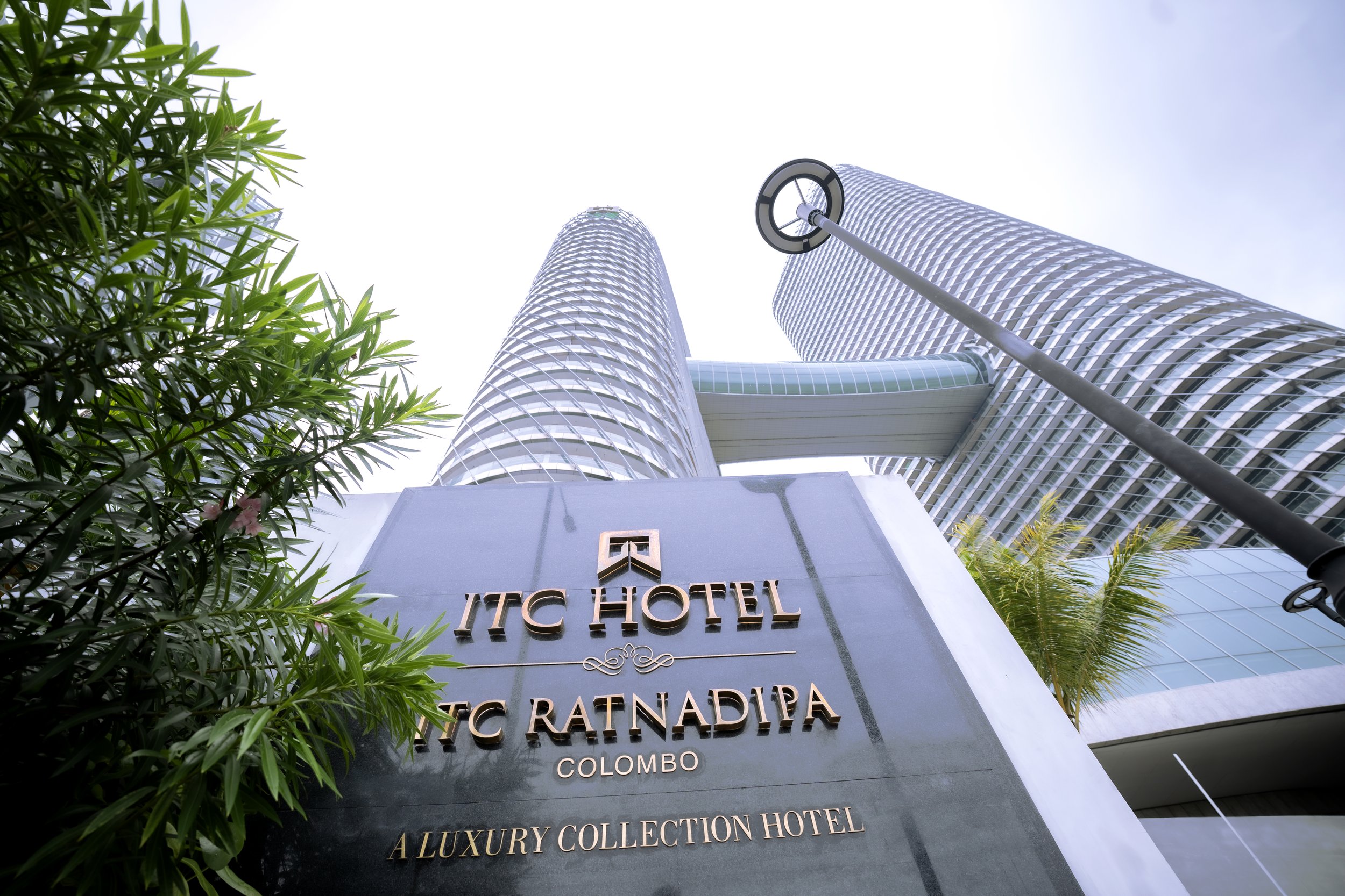 Low-angle view of the ITC Hotel and luxury collection hotel in Colombo, with modern skyscrapers and a street lamp in foreground, greenery on side, under a clear bright sky.