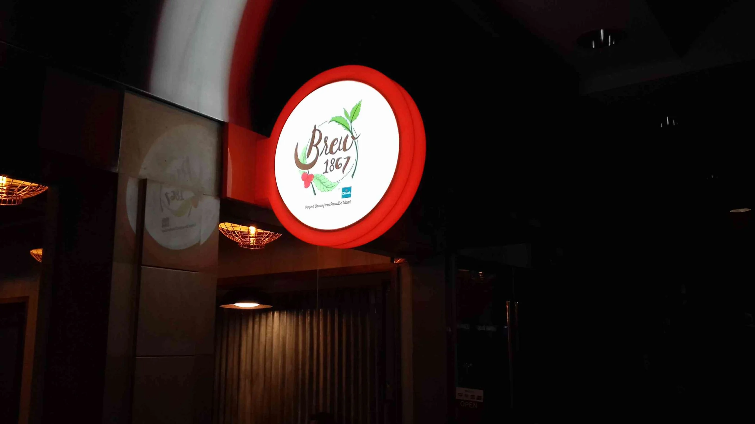 Illuminated circular sign for Brew 1867 with a leaf and flower design, inside a building with warm lighting.