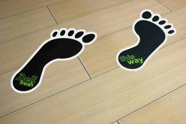 Two black footprints with white borders, one labeled "Deli fresh" and the other labeled "this way" in green, on a wooden floor.