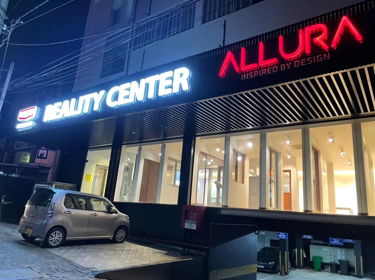Night view of a Beauty Center with illuminated signs for 'Beauty Center' and 'Allura' outside a modern building, with parked cars in front.