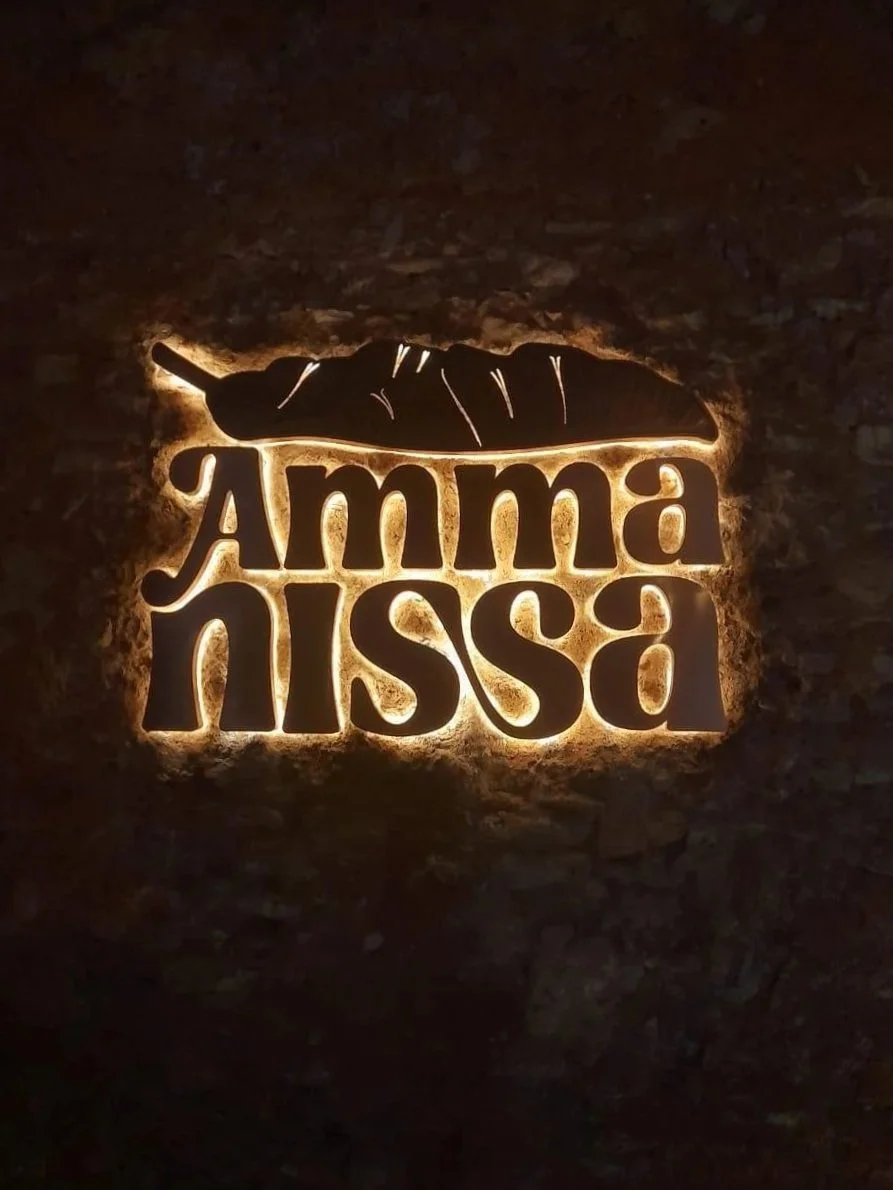 The illuminated sign says 'Amma Nissa' with a graphic of a chili pepper above the text, mounted on a brick wall.