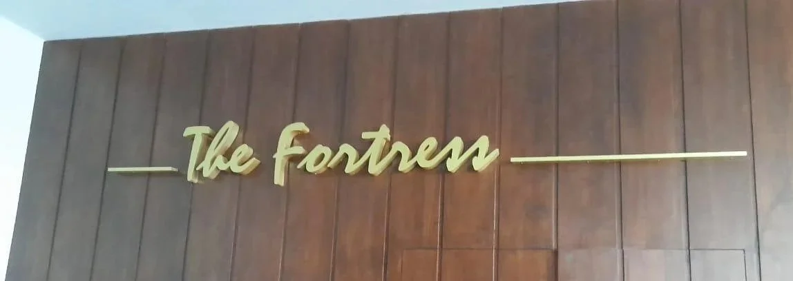 Sign on a wooden wall with the text "The Fortress" in cursive, gold-colored letters.
