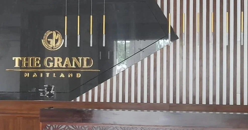 Hotel lobby with black and gold sign reading 'The Grand Maitland,' gold vertical light fixtures, and striped wall design.