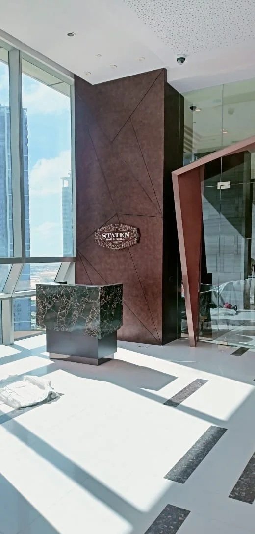 Interior of a modern building with large windows, sunlight casting shadows, a marble-topped reception desk, and a sign for 'Staten Bar & Grill' on a textured wall.