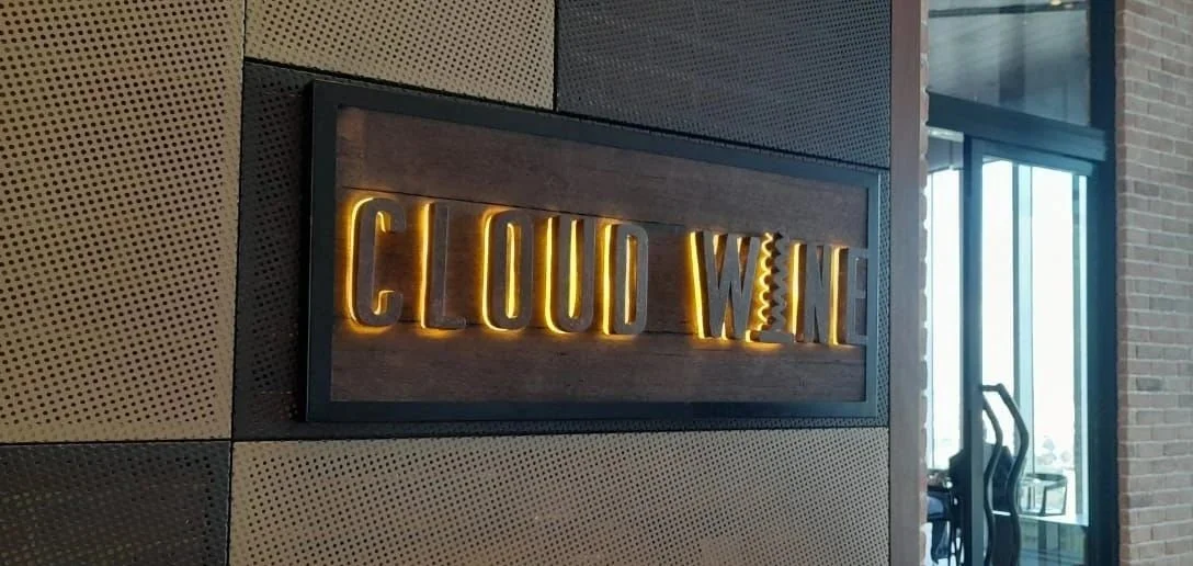 Decorative sign reading "Cloud Wine" with backlit yellow letters, mounted on a wooden panel on a textured wall in a modern interior space.