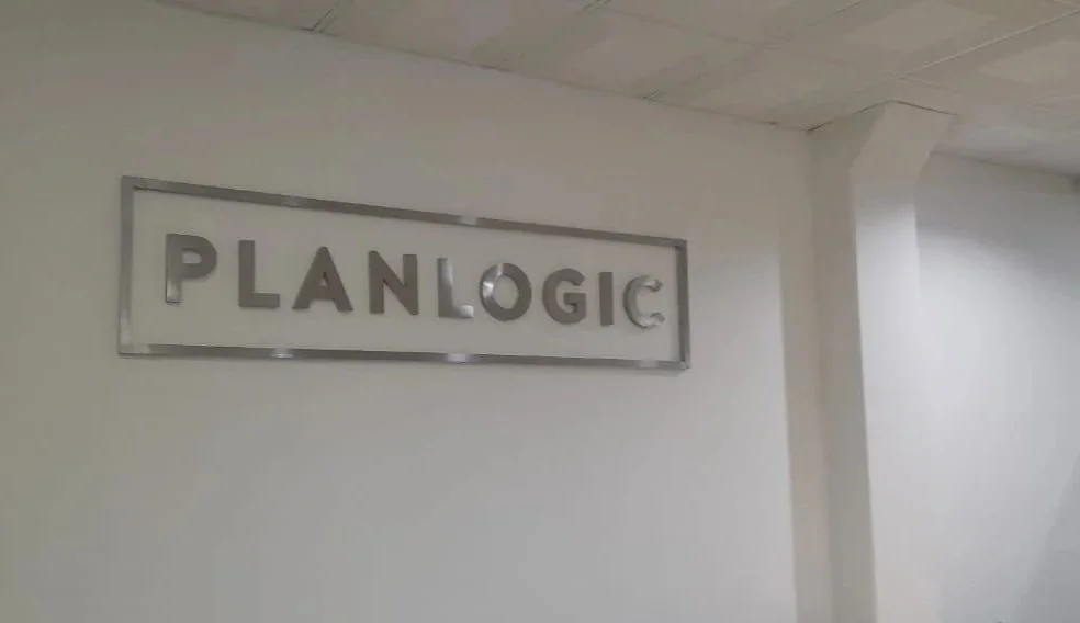 Metal sign reading 'PLAN LOGIC' mounted on a white wall in an office or building interior.