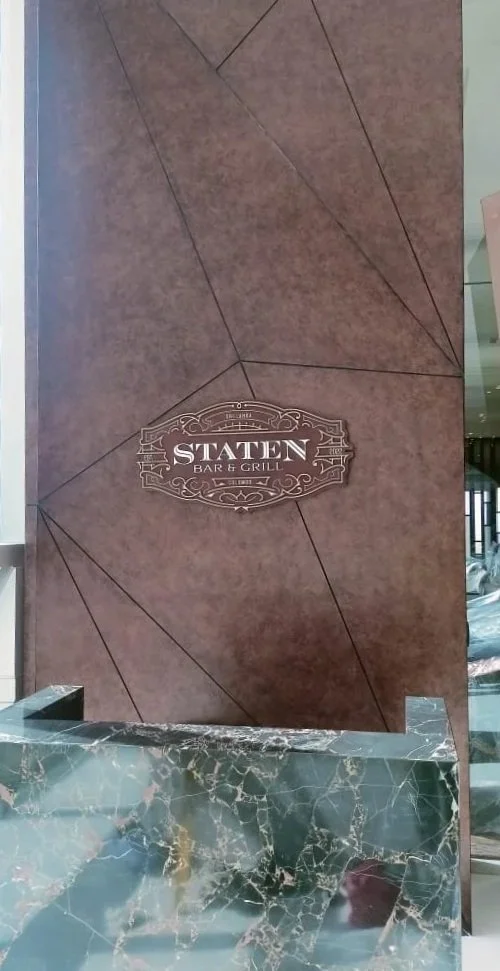 Interior of a restaurant or bar with a brown geometric wall and a marble counter, featuring a sign that reads "Staten Bar & Grill."