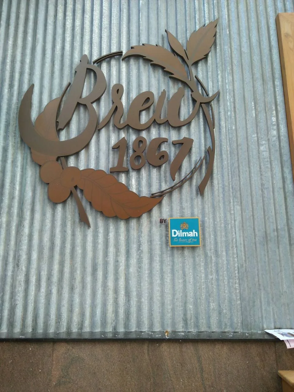 Metal wall decor with the word 'Brew' in cursive, the year 1867, and decorative leaves, mounted on a corrugated metal wall.