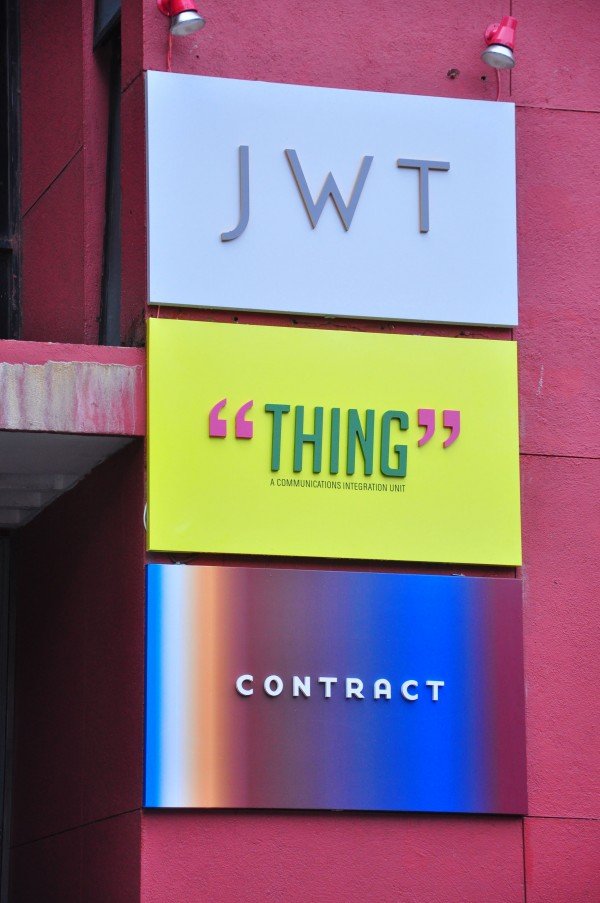 Three signs on a pink building wall. The top sign has the letters "JWT." The middle sign is bright yellow with the word "THING" in bold green and pink quotation marks around it. The bottom sign is a colorful gradient with the word "CONTRACT" in white