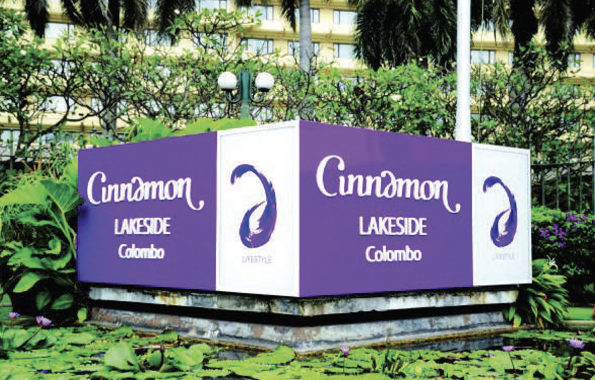 A purple and white sign for Cinnamon Lakeside Colombo, set on a small platform surrounded by water lilies and greenery, with trees and a building in the background.
