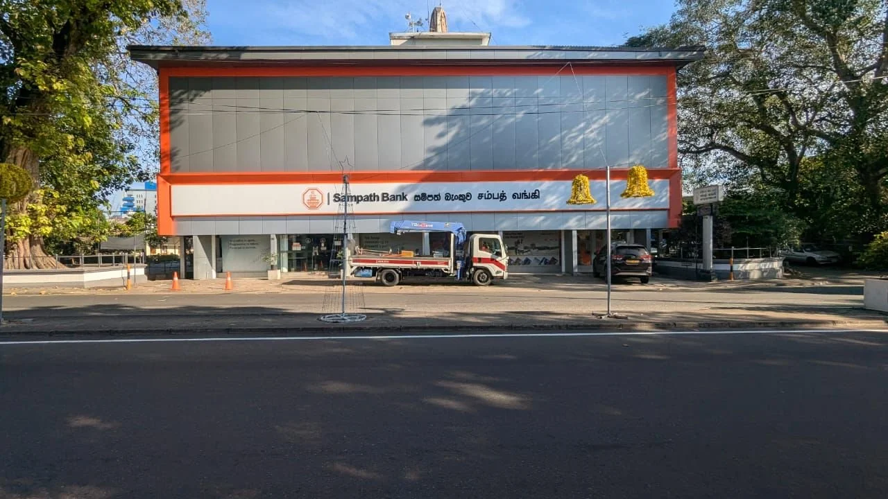 Front view of a Sampath Bank branch building with a truck parked in front and a few cars on the side, surrounded by trees and a clear sky.
