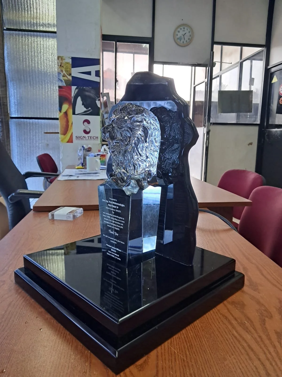A glass lion head sculpture on a black base inside an office or conference room with chairs, a table, and windows in the background.