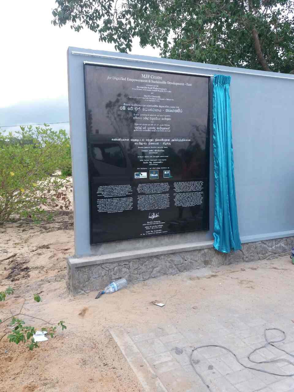 Outdoor display board with black informational sign and blue curtain on right side, surrounded by trees and sandy ground with a water bottle and trash on the ground