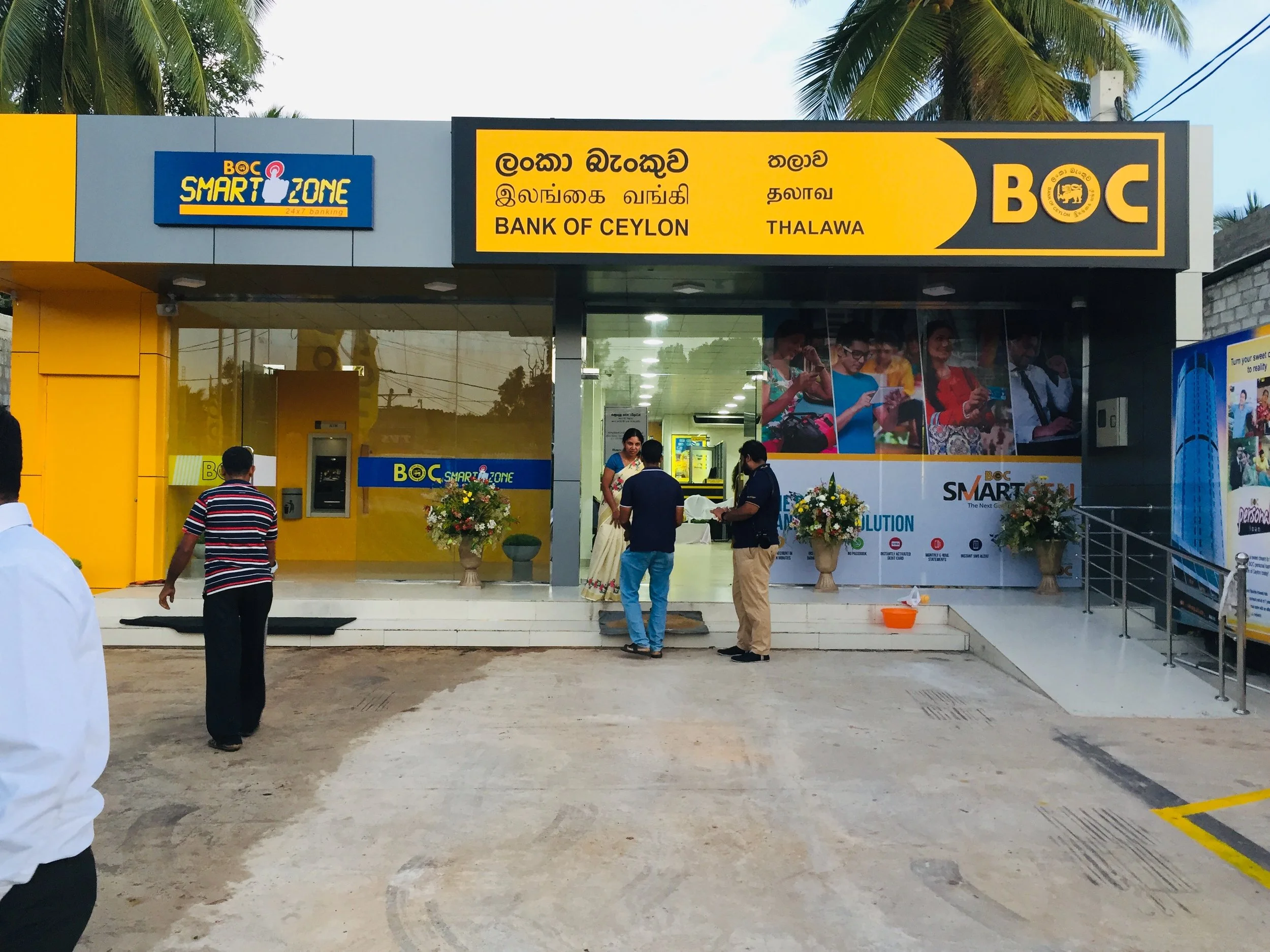 Photo of the exterior of a financial institution, the Bank of Ceylon, with people gathered at the entrance. The building features signs in multiple languages and palm trees in the background.