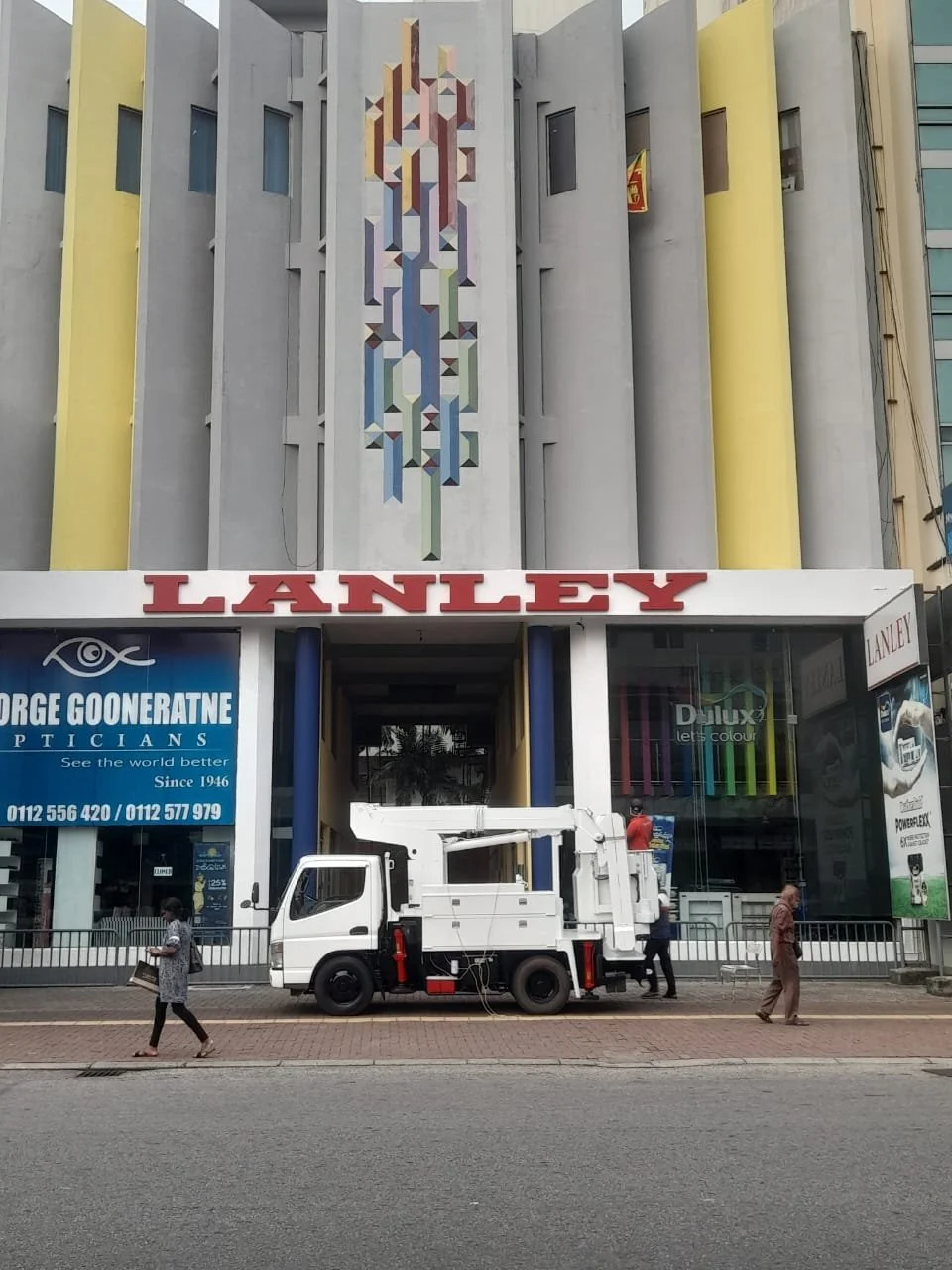 The facade of a building with the name 'LANLEY' in large red letters, featuring architectural details with vertical yellow and gray panels and a central geometric mural. There are shop signs for George Gooneratne Opticians and Dulux Paints. A white u