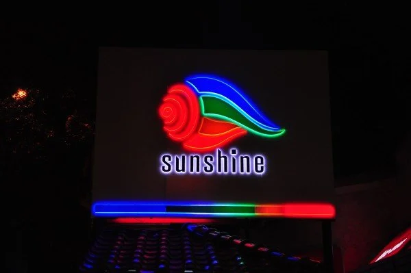Neon sign with colorful snail shell and leaf, with the word 'Sunshine' beneath it, on a dark background.