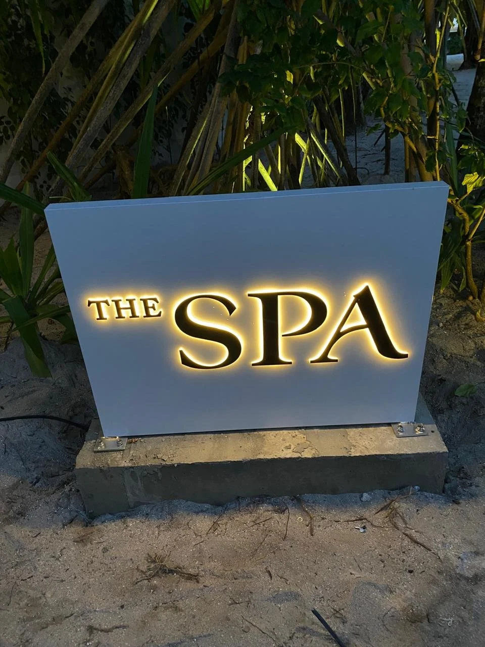 A sign illuminated with yellow backlighting reads 'The SPA'. The sign is placed on a concrete base surrounded by sand and greenery.