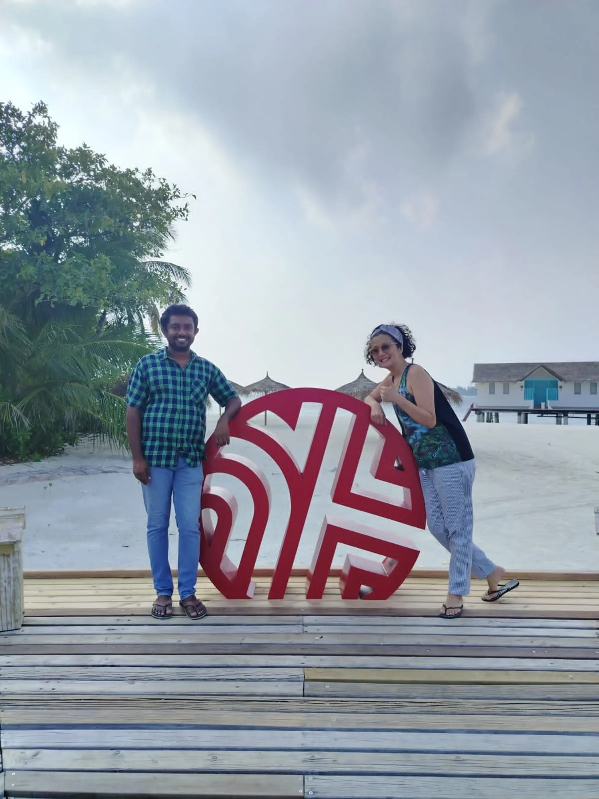 A man and a woman standing on a beach with overwater bungalows in the background, holding a large red and white 'SOY' sign, surrounded by tropical trees.