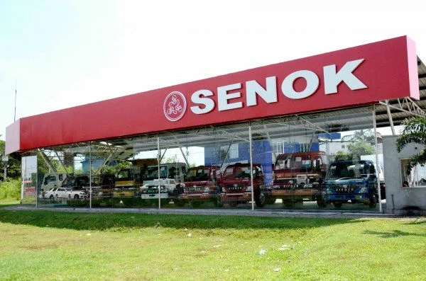 Exterior of a vehicle showroom with a red sign that says "SENOK" and glass walls displaying trucks and cars inside.