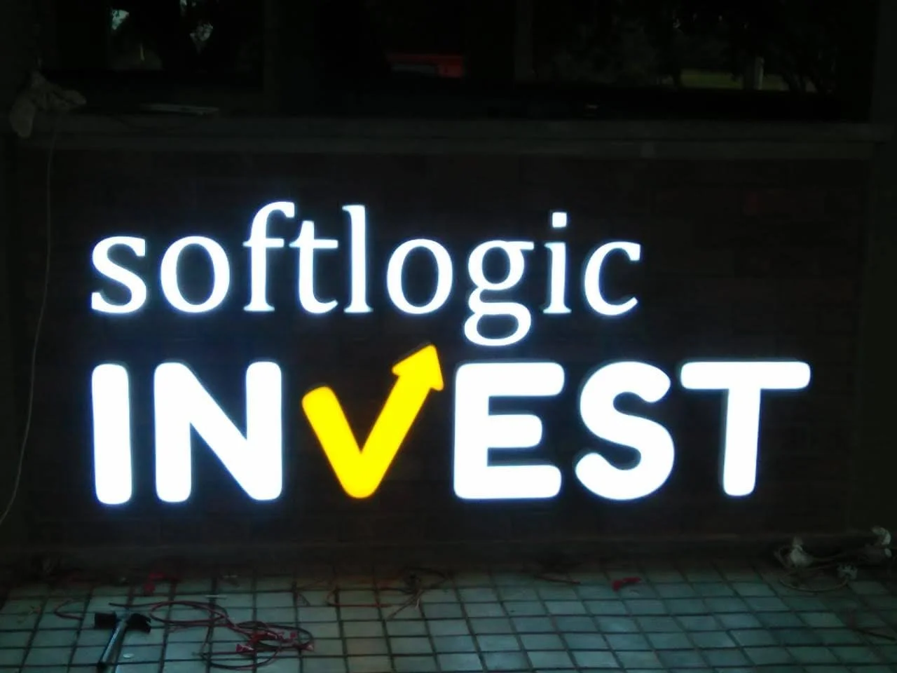 Illuminated sign with the words 'softlogic INVEST', where the 'V' is styled as an upward arrow in yellow.