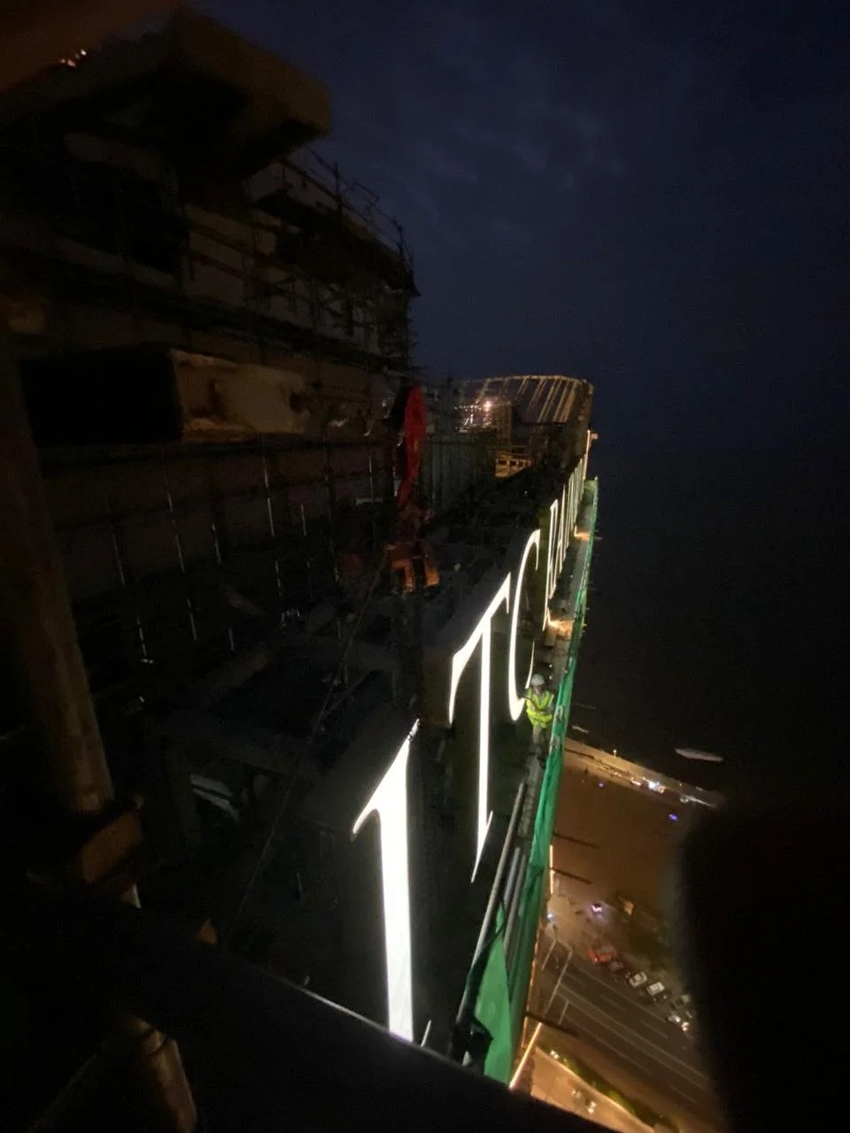 Nighttime photo of large building signage displaying the number '700', with construction workers working on the sign and scaffolding overhead, and a parking lot below.