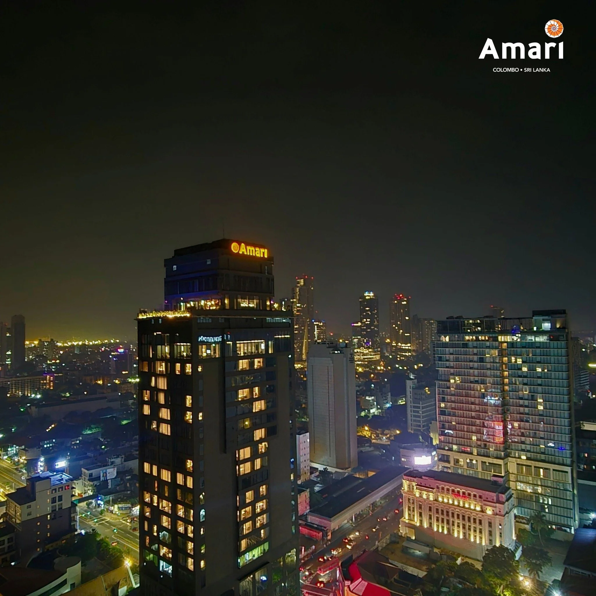 Nighttime cityscape in Colombo, Sri Lanka, featuring illuminated high-rise buildings, streets with moving traffic, and the Amari hotel sign visible at the top of a tall building.