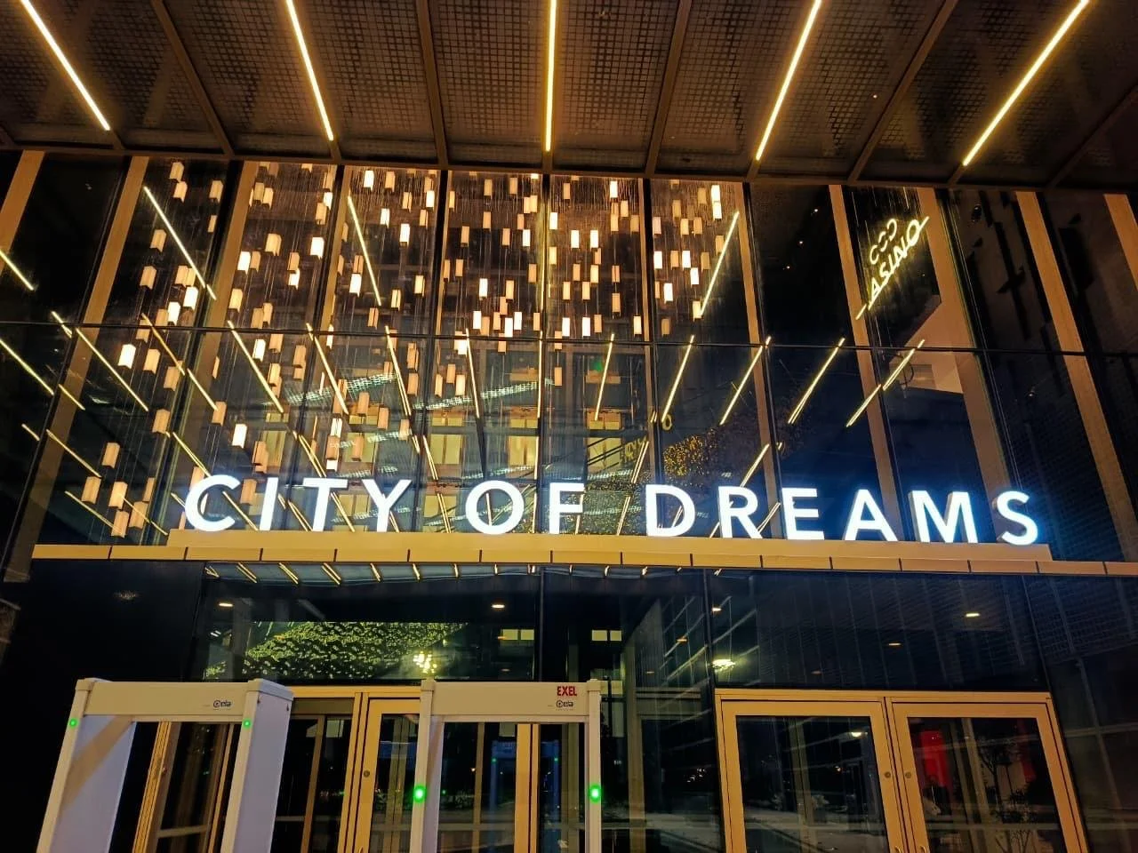 Night view of the modern facade of City of Dreams building with illuminated sign, glass windows, decorative lighting, and a security gate at the entrance.