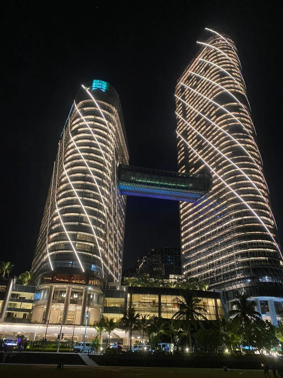 Night view of two illuminated skyscrapers connected by a skybridge, with palm trees and a busy area at the base.