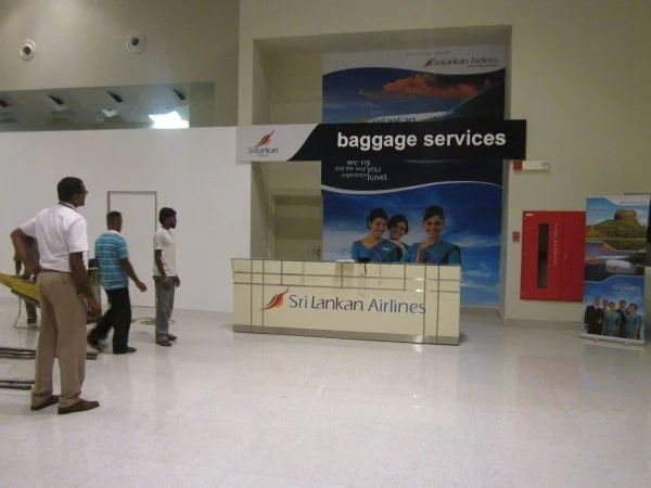 Passengers at Sri Lankan Airlines baggage services counter in an airport, with three people walking nearby and a red fire hose reel on the wall.