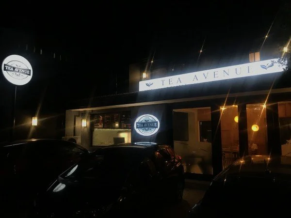 Nighttime exterior of a building with illuminated signs reading 'Tea Avenue' and 'Tea & Era,' with parked cars in front.