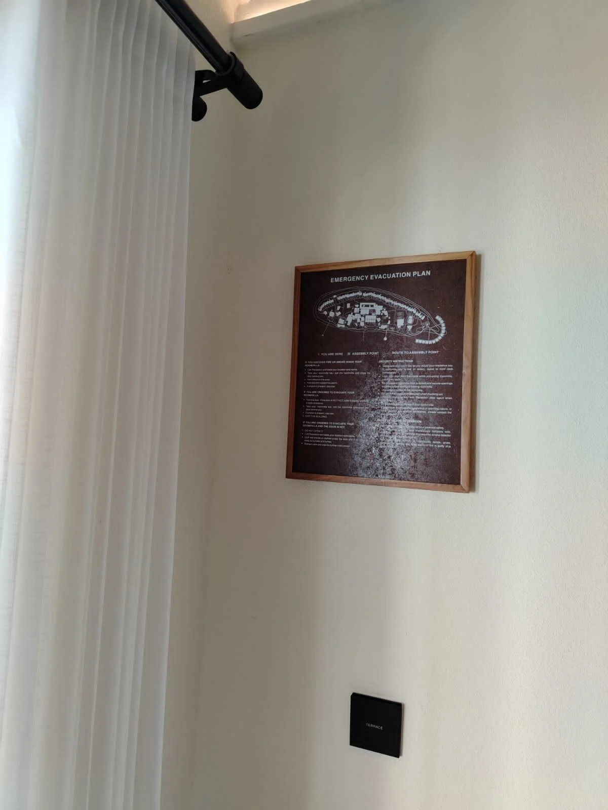 A framed emergency evacuation plan on a light-colored wall, with vertical blinds on the left and a black thermostat below.