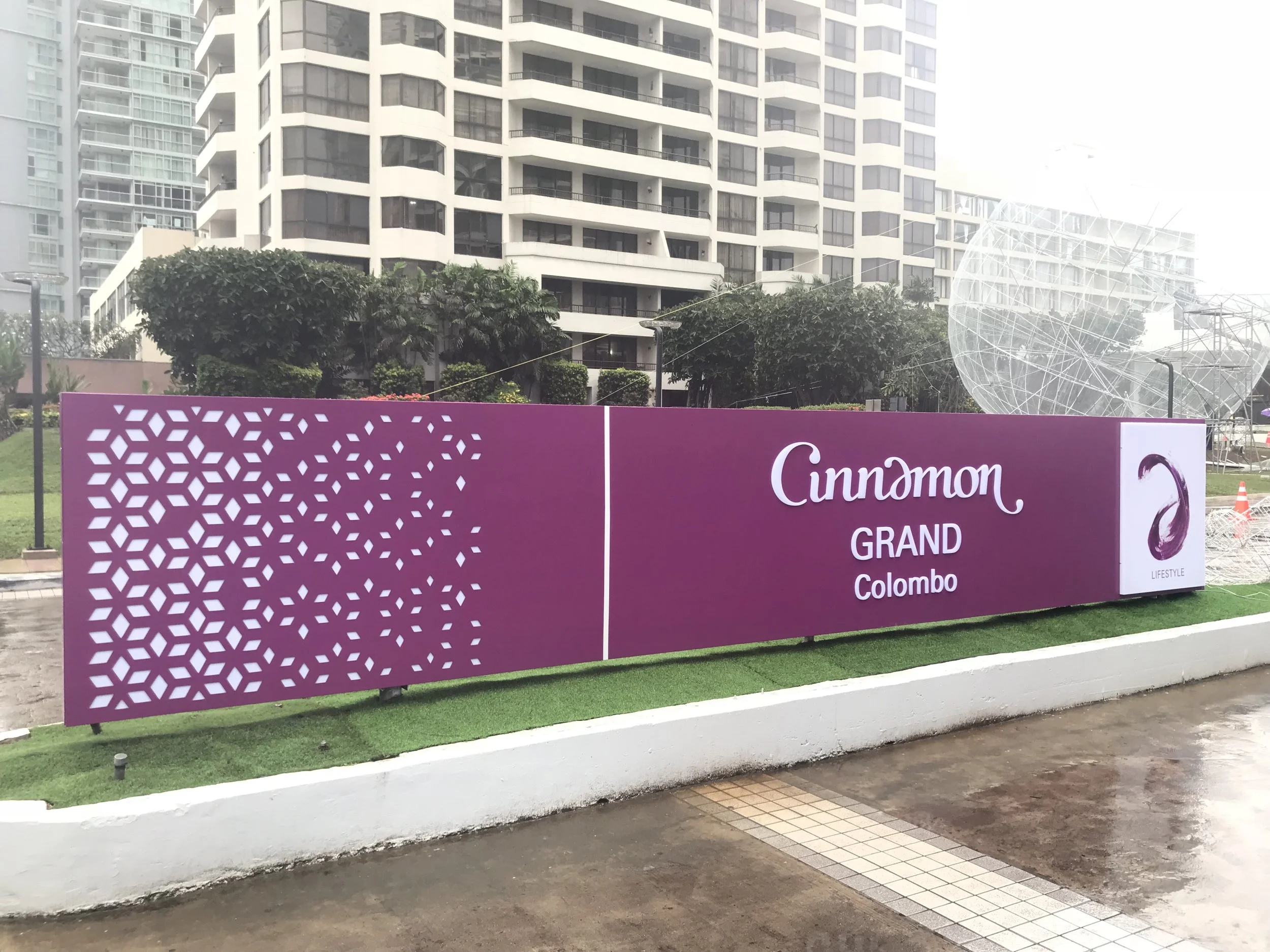 A large purple sign with white decorative patterns and text that reads "Cinnabon GRAND Colombo" along with a logo, situated in front of high-rise residential buildings and some greenery in an urban area.
