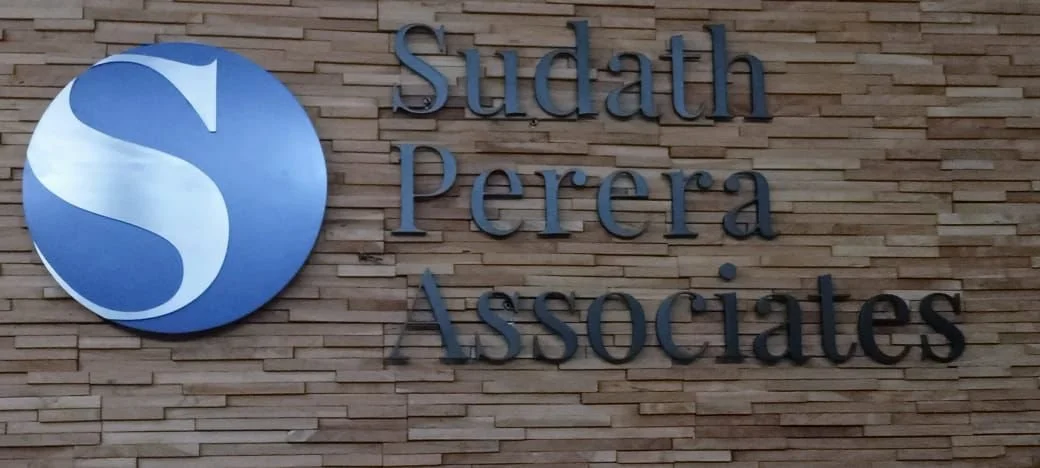 Sign for Studath Perera & Associates on a brick wall, with a circular blue logo featuring a white stylized 'S'.