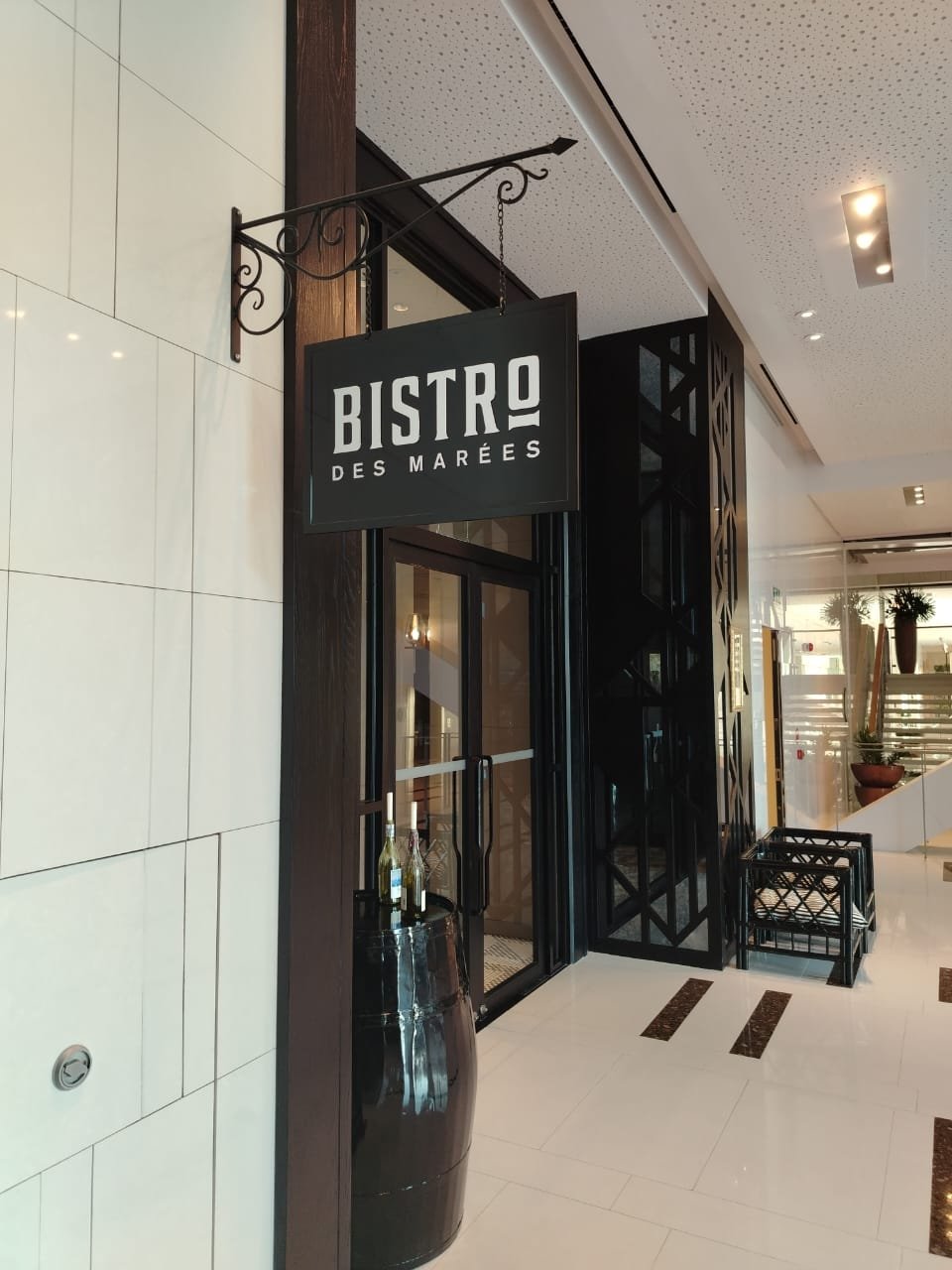 Restaurant entrance with a black sign that reads 'Bistro des Marées,' decorative black screen and a black barrel with bottles outside.