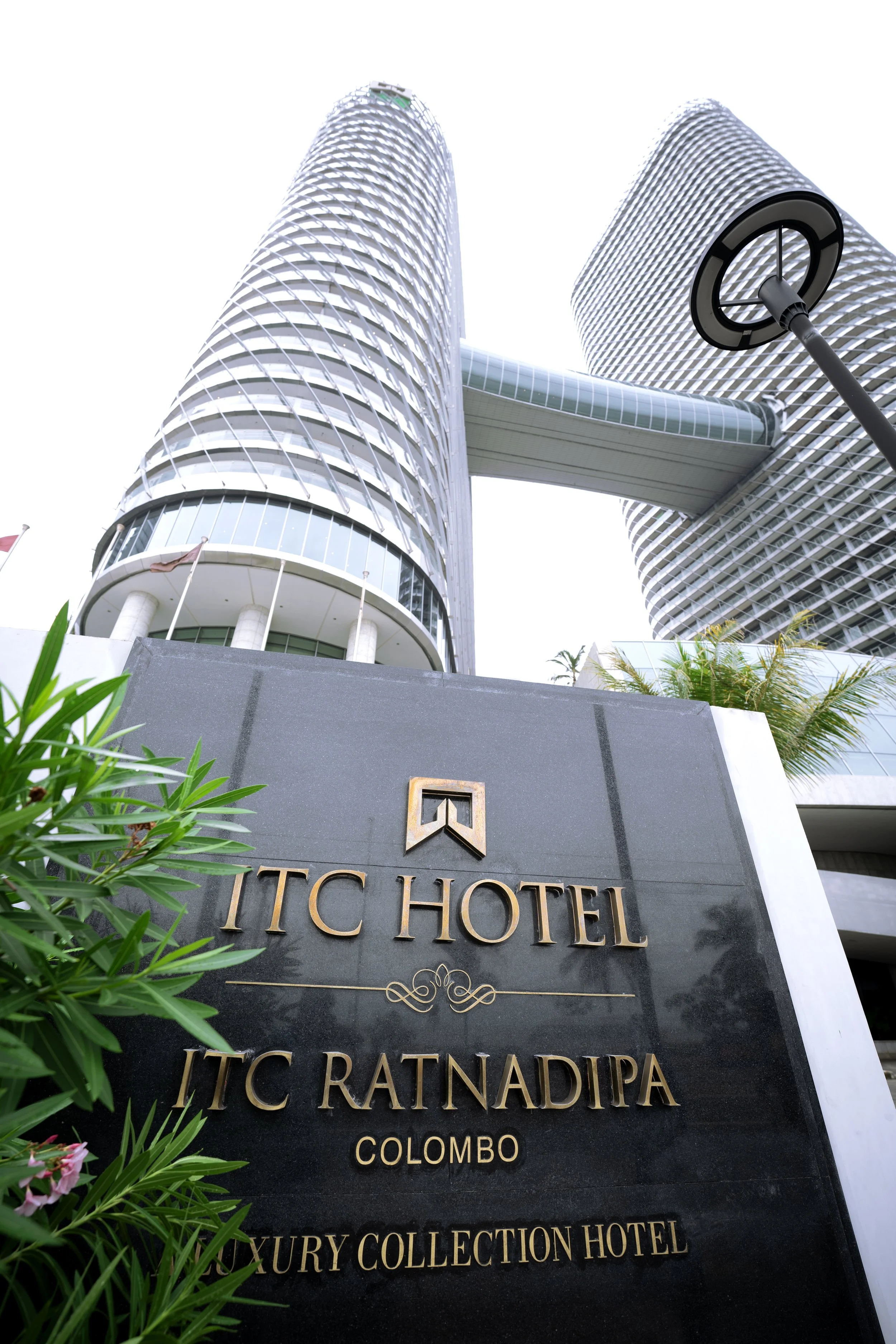 View of ITC Ratanapira luxury hotel in Colombo, featuring modern skyscrapers with glass facades and connecting skywalks, with greenery in the foreground and a streetlamp.
