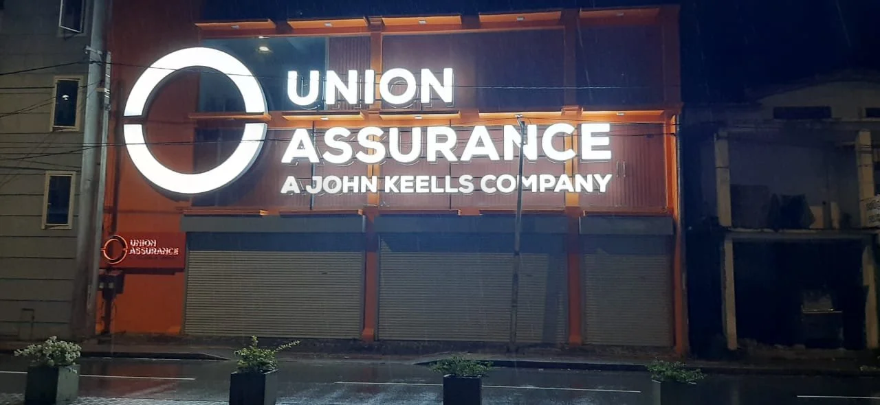 Night view of a large, illuminated sign for Union Assurance, a John Keells company, on a building with closed rolling shutters.